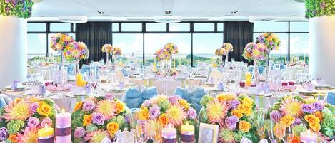 Un salón de banquetes con mesas redondas, centros de mesa florales y vistas amplias a través de grandes ventanales.