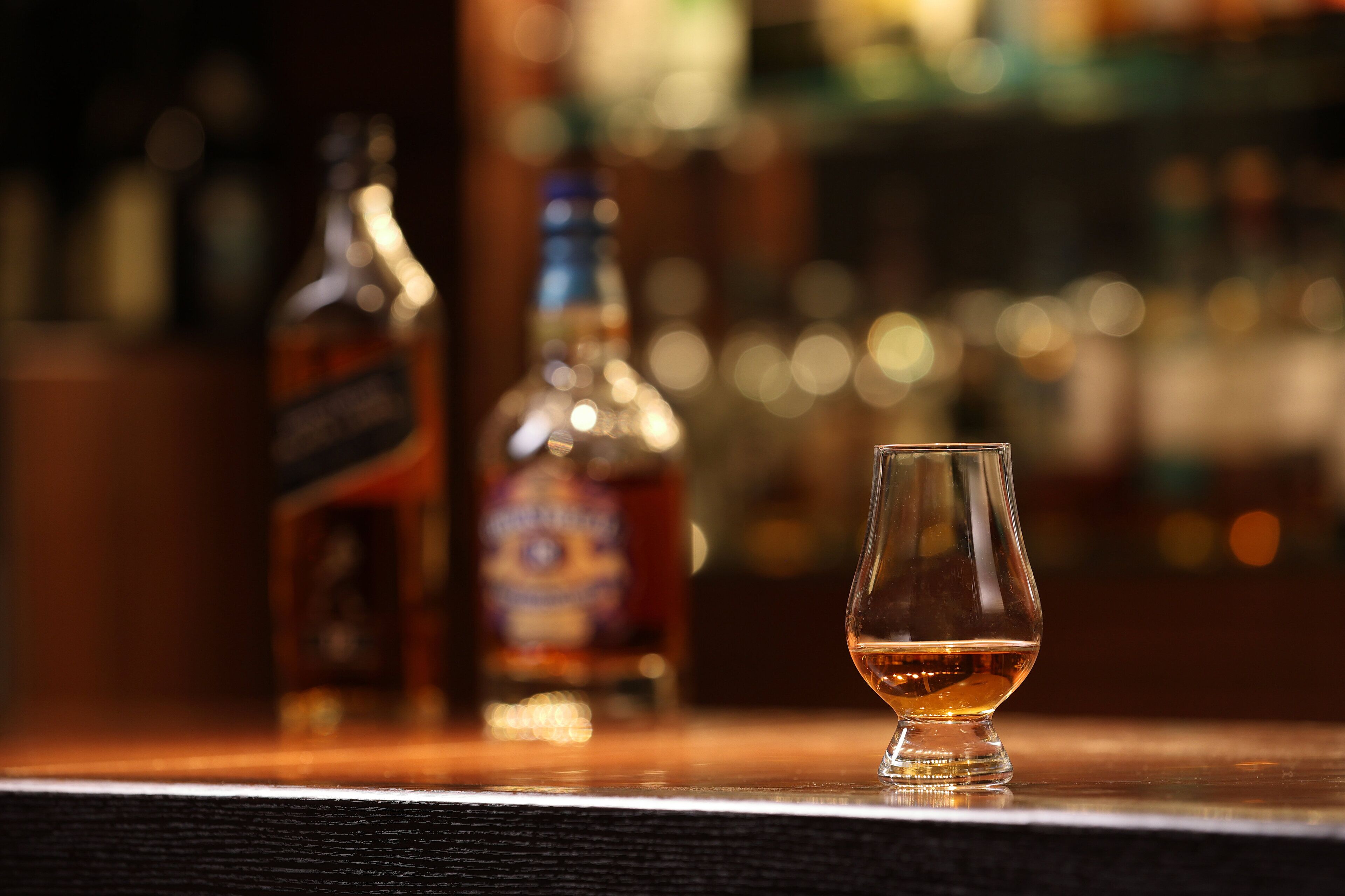A bar setting with a glass of whiskey and two bottles in the background.