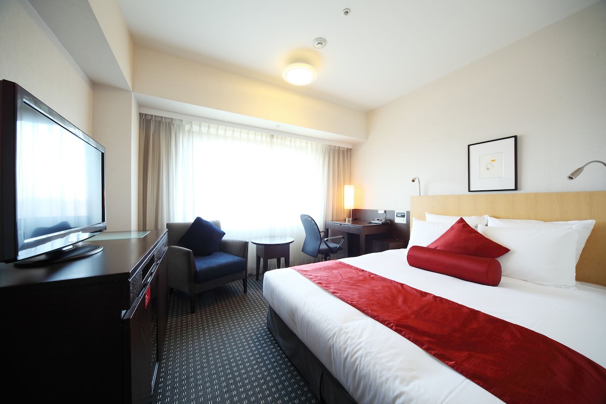 Een hotelkamer met een bed, een bureau, een televisie, een stoel en een raam met gordijnen.