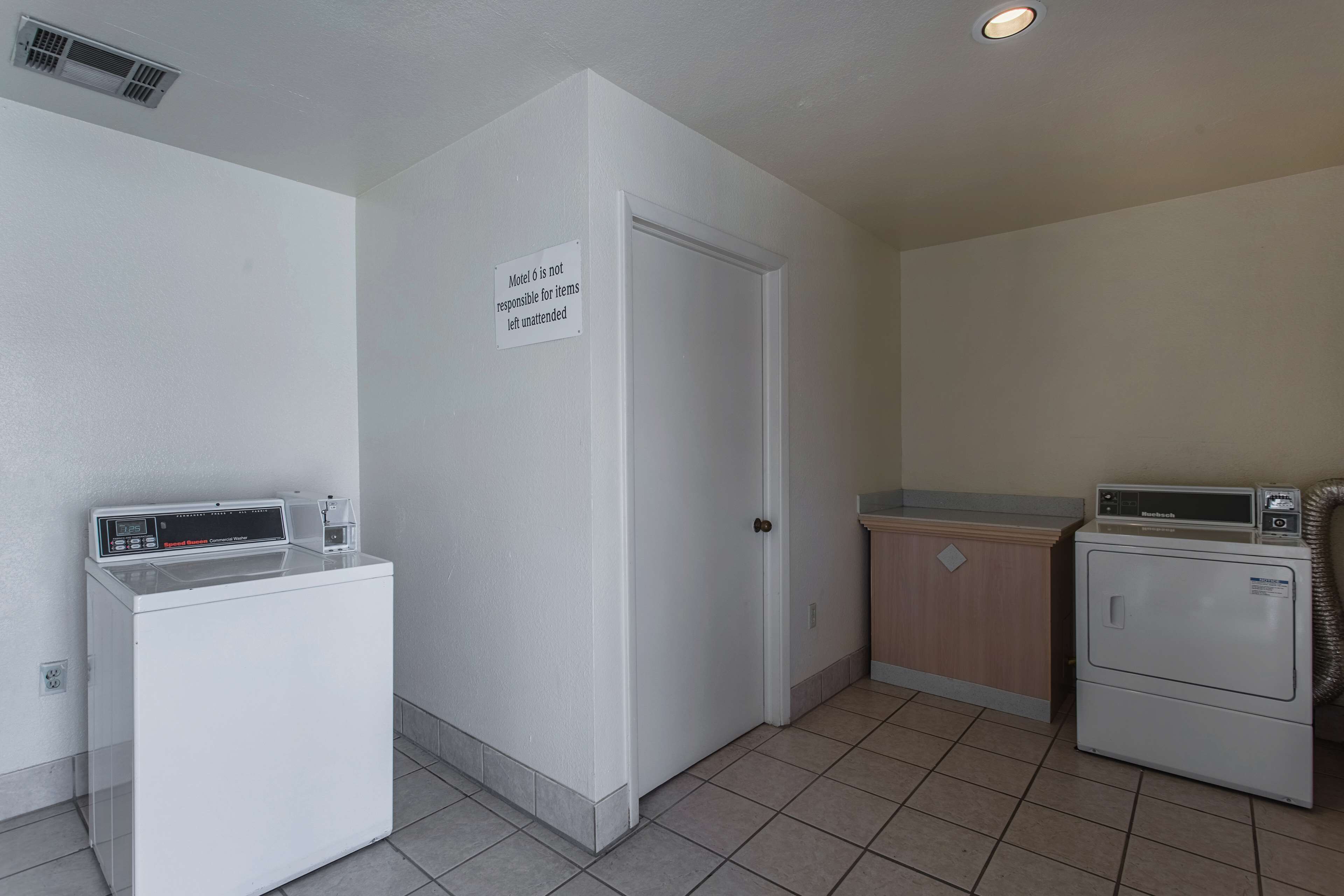 A laundry room with a front-loading washer and dryer, a sign stating 'Motel is not responsible for items left unattended', and a door leading to another room.