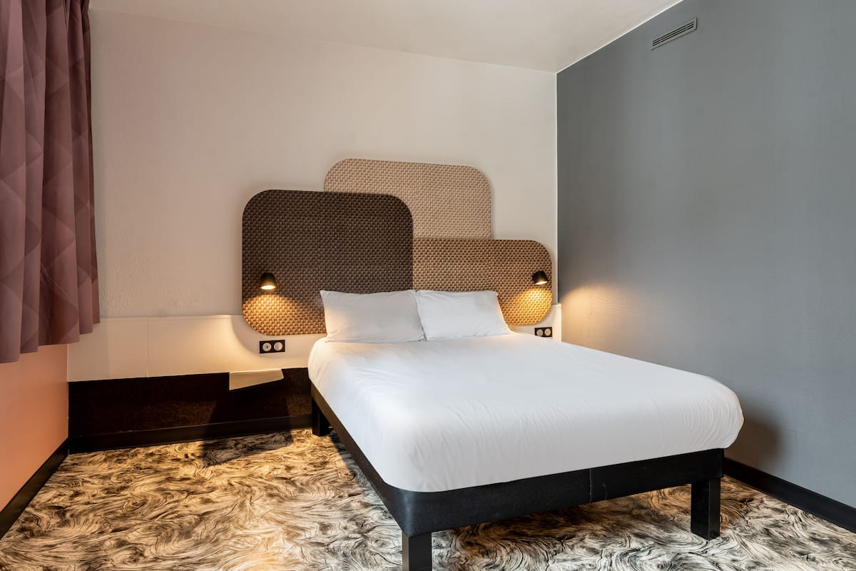 Une chambre d’hôtel moderne, dotée d’un lit, d’une tête de lit avec éclairage intégré, d’une moquette à motifs et d’un banc de couleur sombre.