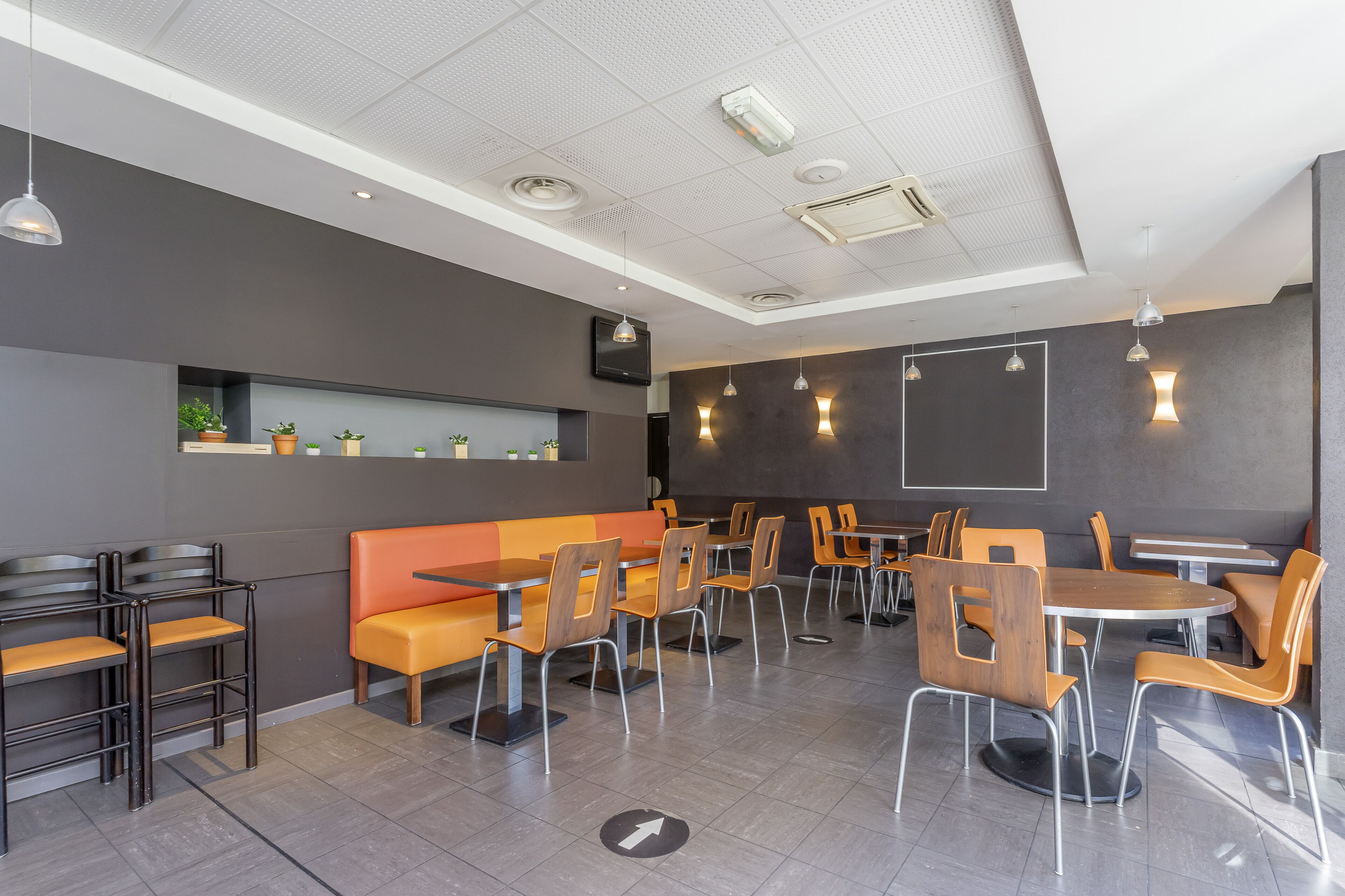 Un restaurante moderno con mesas y sillas altas, una barra larga y una zona de estar con cojines naranjas.
