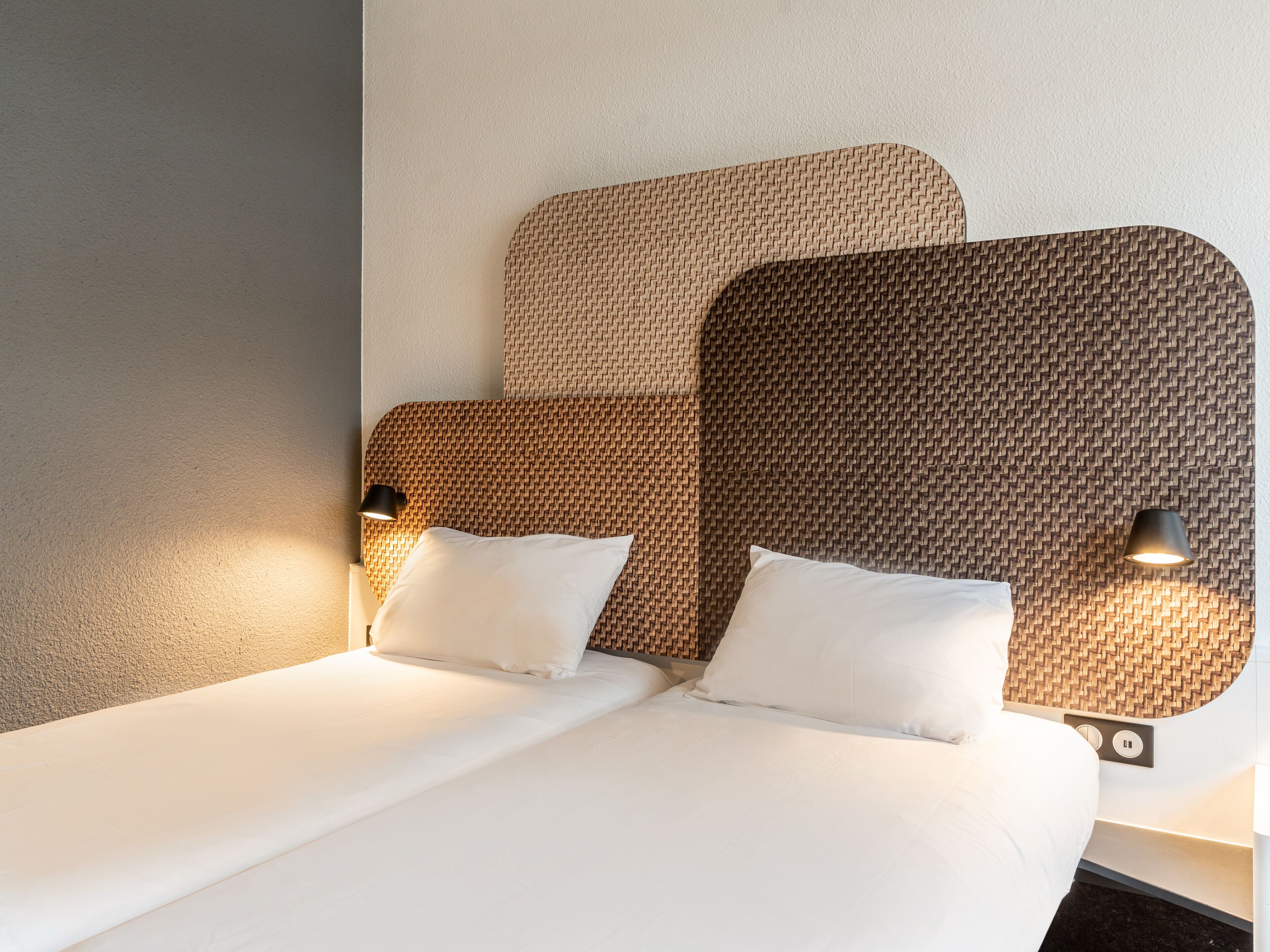A neatly made bed with white linens and two pillows, a headboard with a woven pattern, and two wall-mounted lamps on either side.