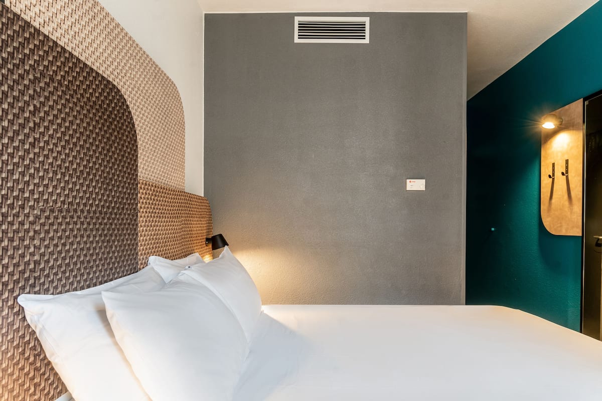 Une chambre d’hôtel moderne avec un lit, une tête de lit en osier, un mur gris, une grille d’aération et un luminaire fixé au mur.