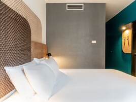 Une chambre d’hôtel moderne avec un lit, une tête de lit en osier, un mur gris, une grille d’aération et un luminaire fixé au mur.