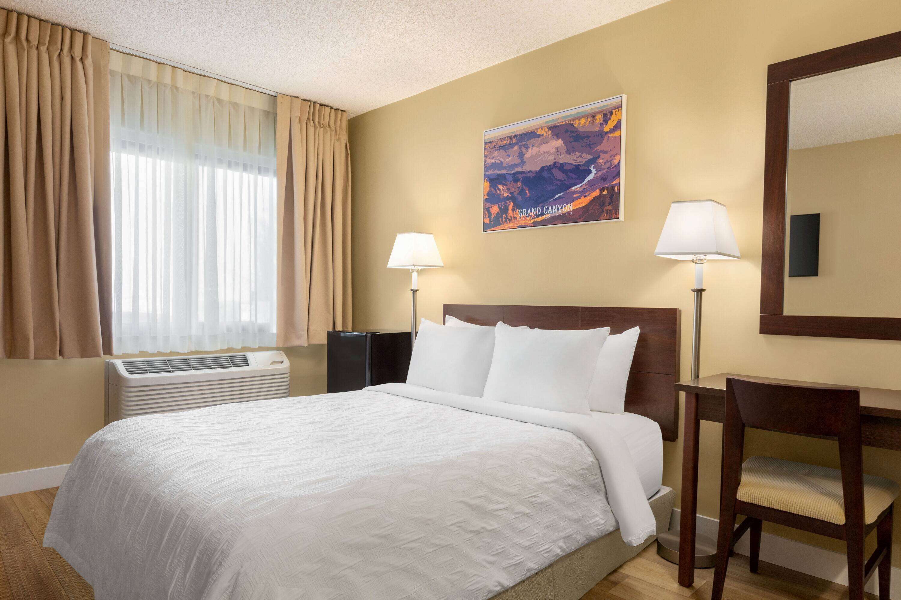 A hotel room with a large bed, two bedside lamps, a desk with a chair, a window with curtains, an air conditioner, and a wall-mounted painting of the Grand Canyon.