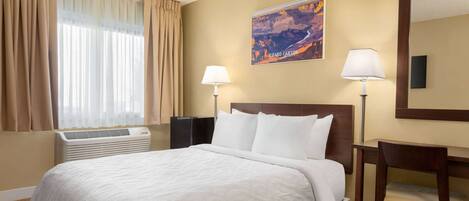 A hotel room with a large bed, two bedside lamps, a desk with a chair, a window with curtains, an air conditioner, and a wall-mounted painting of the Grand Canyon.