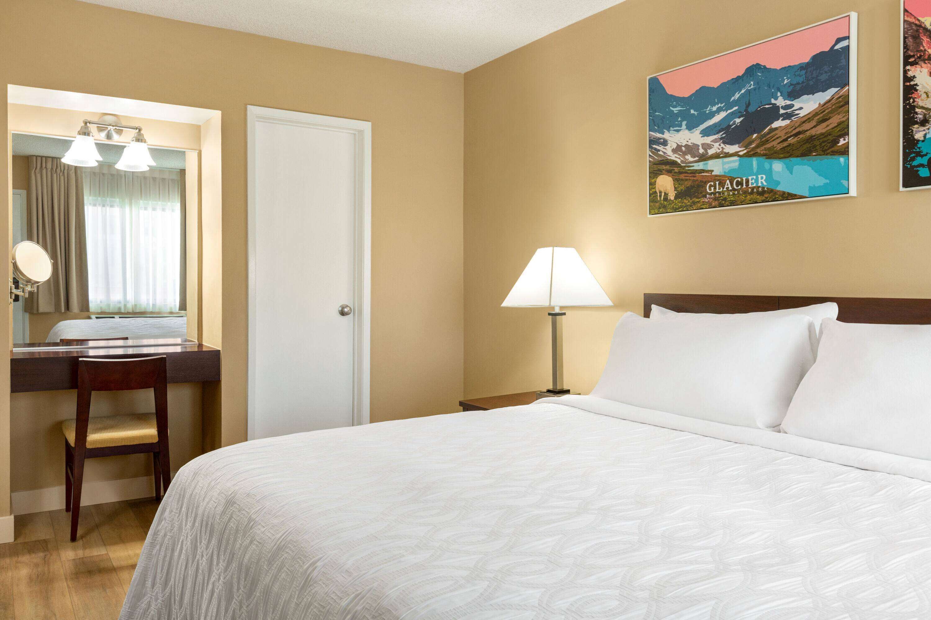 A hotel room with a bed, a desk with a chair, a lamp, and a painting of a glacier.