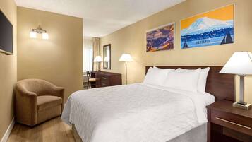 A hotel room with a large bed, a brown armchair, a desk, and two wall-mounted lamps. There are two framed pictures on the wall, one depicting a mountain and the other the Olympic Games.