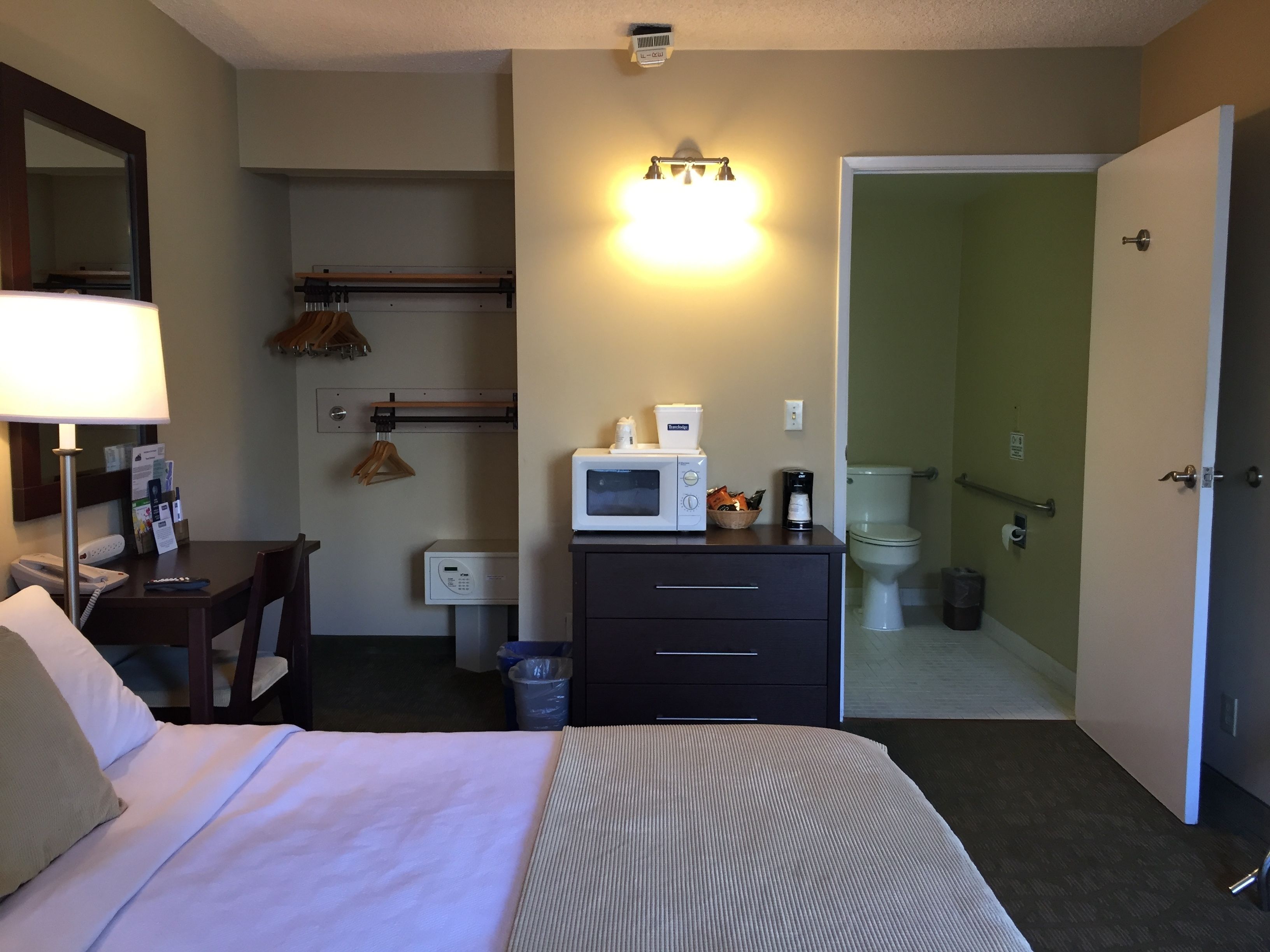 Habitación de hotel con cama, mesita de noche, lámpara, microondas, cafetera y un armario pequeño.