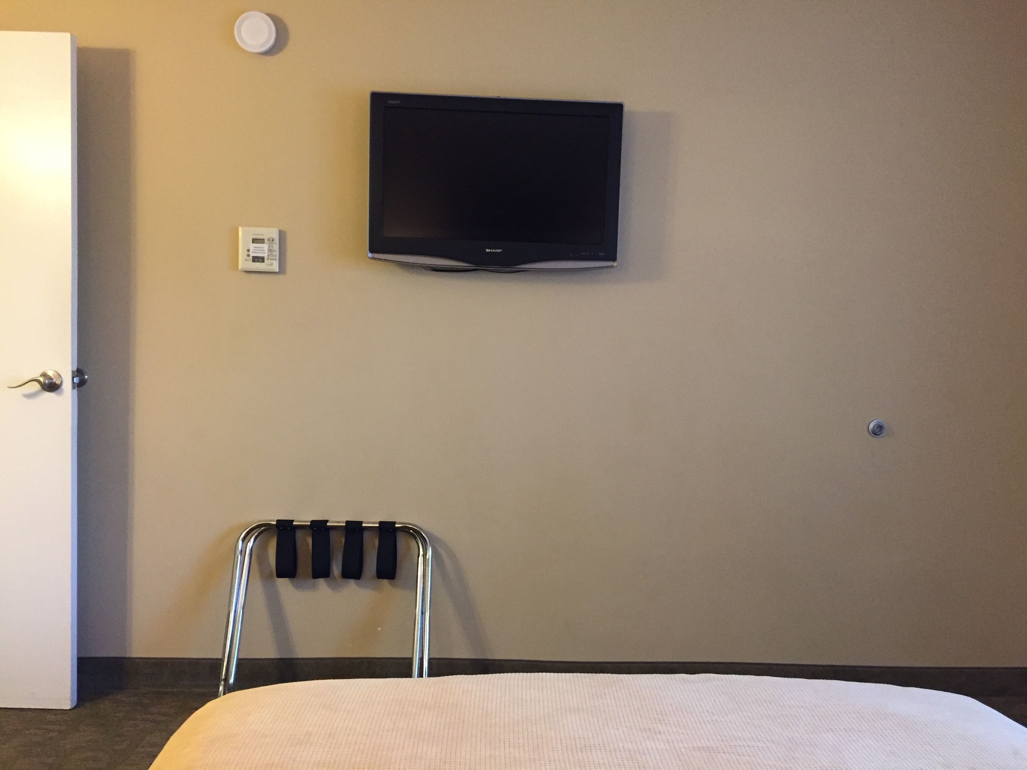 Quarto de hotel com cama, uma cadeira, televisão presa na parede e uma porta.
