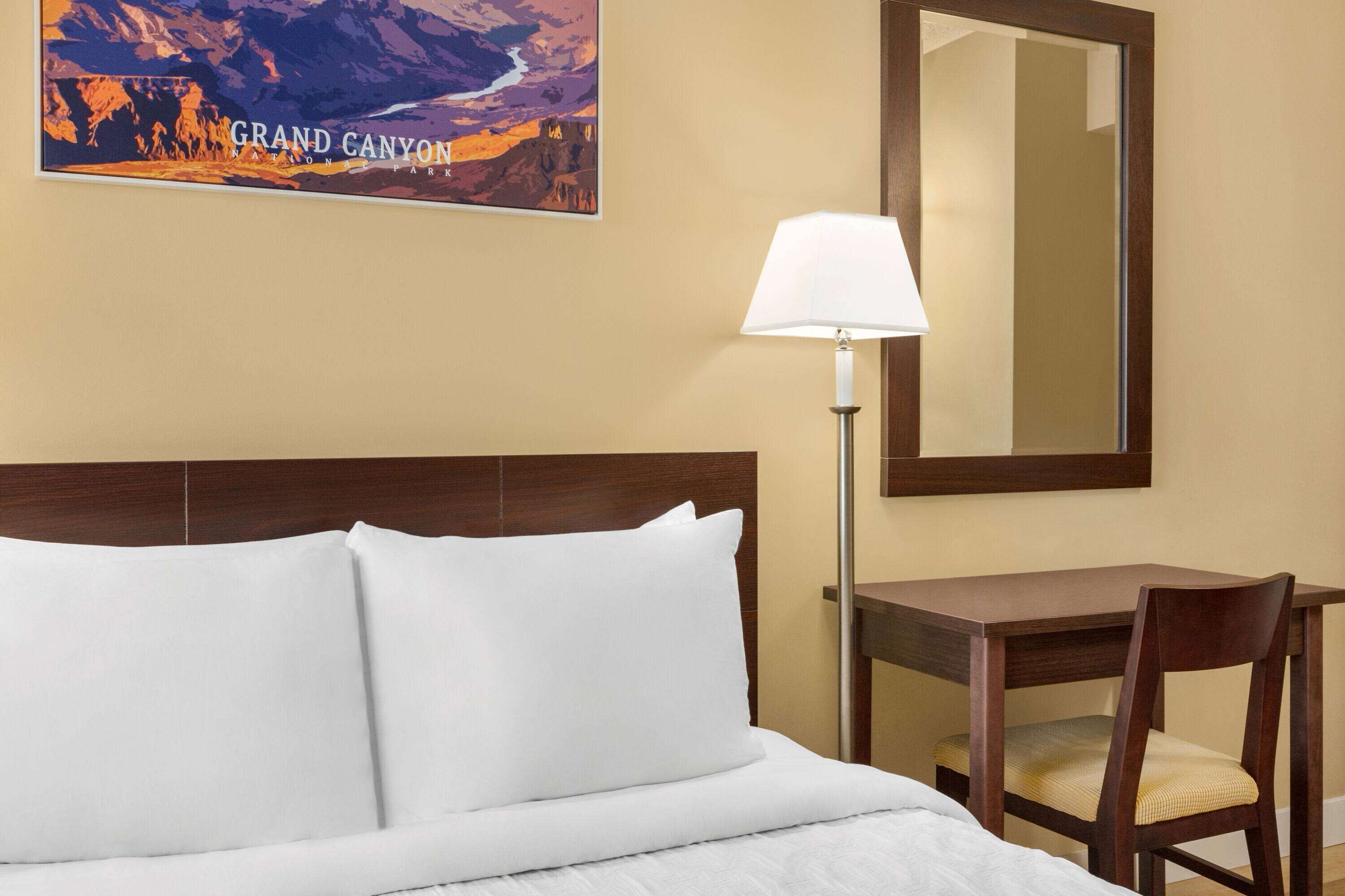 A hotel room with a bed, a desk, a chair, a lamp, and a poster of the Grand Canyon.