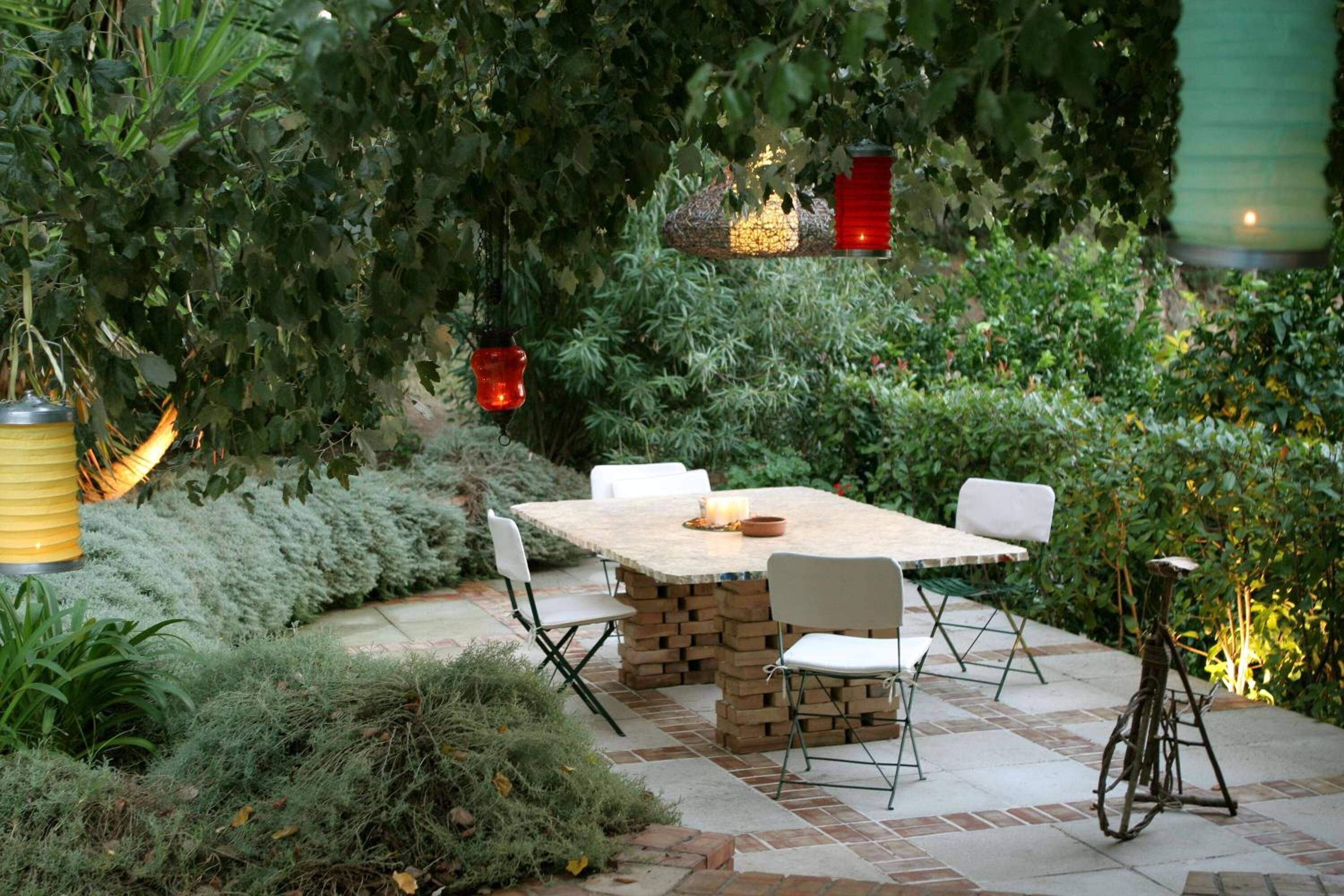 Un patio al aire libre con una mesa de ladrillo y sillas blancas, rodeado de abundante vegetación y faroles decorativos.