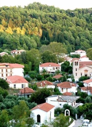 Un pueblo en una ladera con arquitectura tradicional, incluyendo edificios con techos de tejas rojas y paredes blancas, rodeado de frondosos árboles verdes.