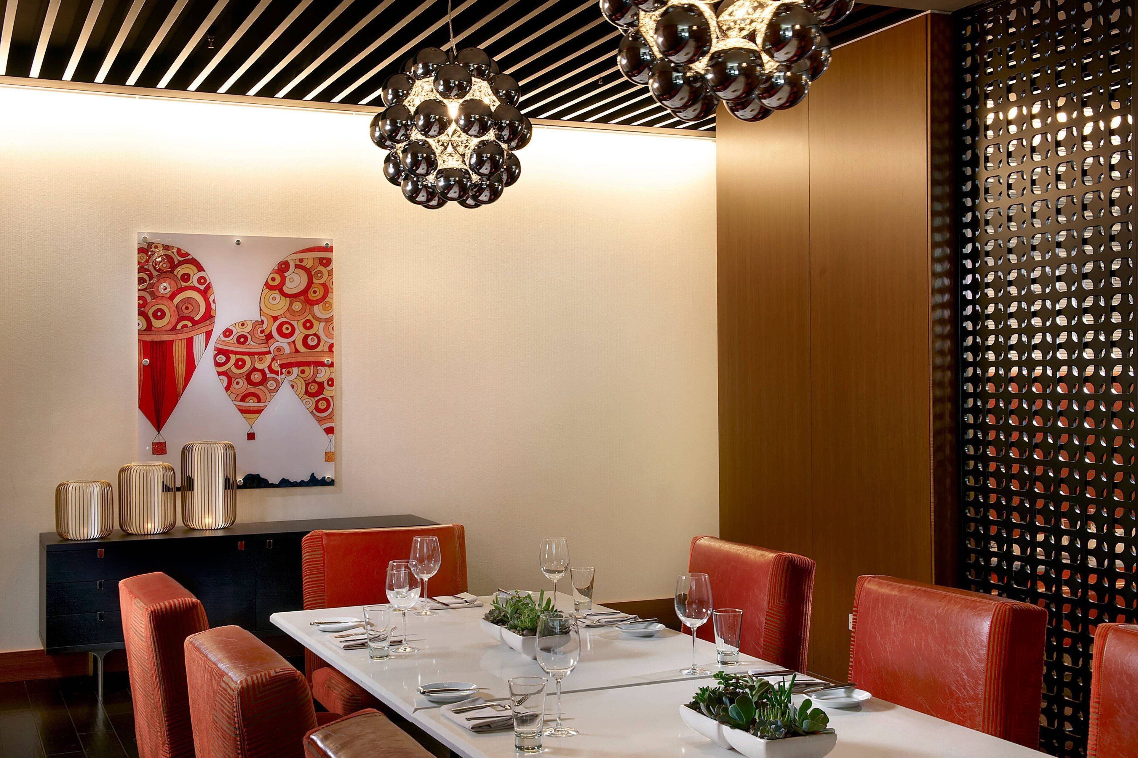 A dining area with a long table set for four, red upholstered chairs, a decorative wall with a patterned artwork, and a ceiling with a unique spherical light fixture.