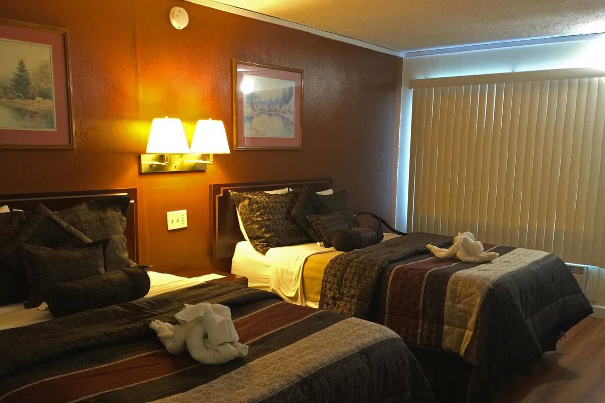 A hotel room with two beds, each with dark bedding and decorative pillows. There are wall-mounted lamps and framed pictures on the walls.