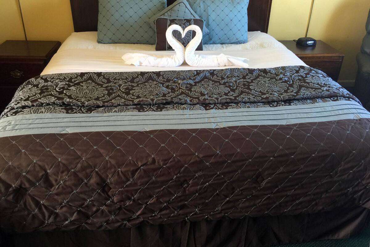 A neatly made bed with a brown quilted bedspread, a decorative pillow with a heart-shaped towel, and a bedside table with a lamp.