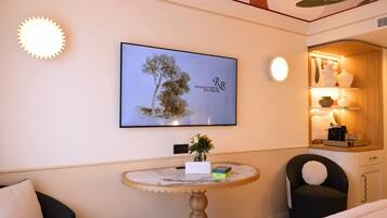 A hotel room with a bed, a round table with a vase and a box, a chair, a television mounted on the wall, and a decorative ceiling.