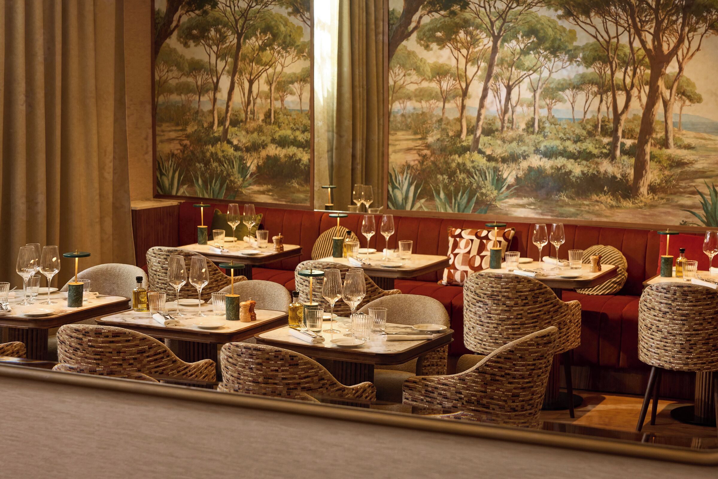 Ein Restaurant mit Korbstühlen, Tischen, die mit Weingläsern und Tellern gedeckt sind, und einem großen Landschaftsbild an der Wand.