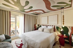 A hotel room with a large bed, a seating area with a chair and table, a balcony with curtains, and a ceiling with abstract art. - Grand Hôtel Roi René Aix - MGallery Collection (Aix-en-Provence)
