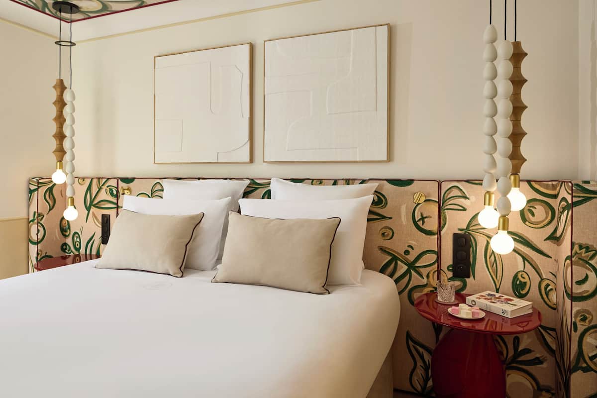 A neatly made bed with white linens, a headboard with a leaf pattern, two framed artworks on the wall, and a bedside table with a lamp and a book.
