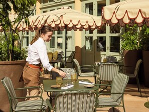 An outdoor dining area with wicker chairs and tables, a waiter serving, and a building with large windows in the background. - Grand Hôtel Roi René Aix - MGallery Collection (Aix-en-Provence)