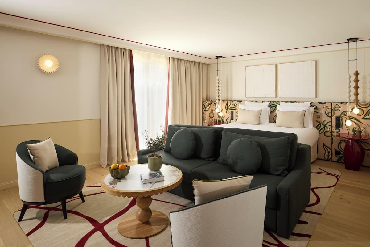 A hotel room with a bed, a green sofa, a round coffee table, a chair, and a small table with a plant and fruit.