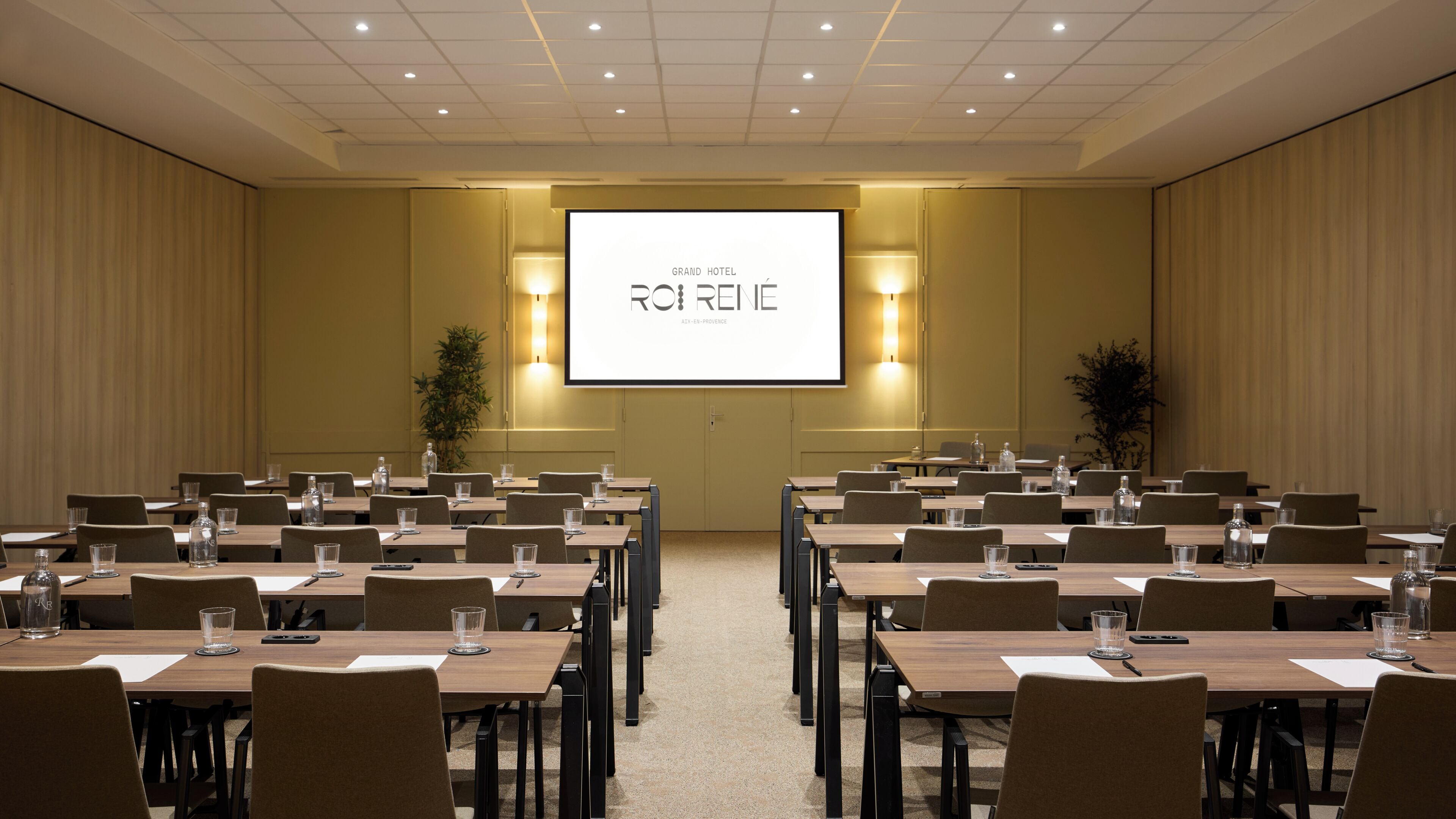 Un salón de conferencias con varias mesas y sillas, una pantalla grande mostrando 'GRAND HOTEL ROIRENÉ' y dos lámparas fijadas a la pared.