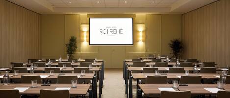 Un salón de conferencias con varias mesas y sillas, una pantalla grande mostrando 'GRAND HOTEL ROIRENÉ' y dos lámparas fijadas a la pared.