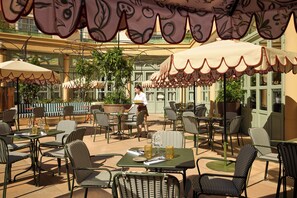 An outdoor patio with wicker chairs and tables, covered by patterned umbrellas, and a waiter walking by.