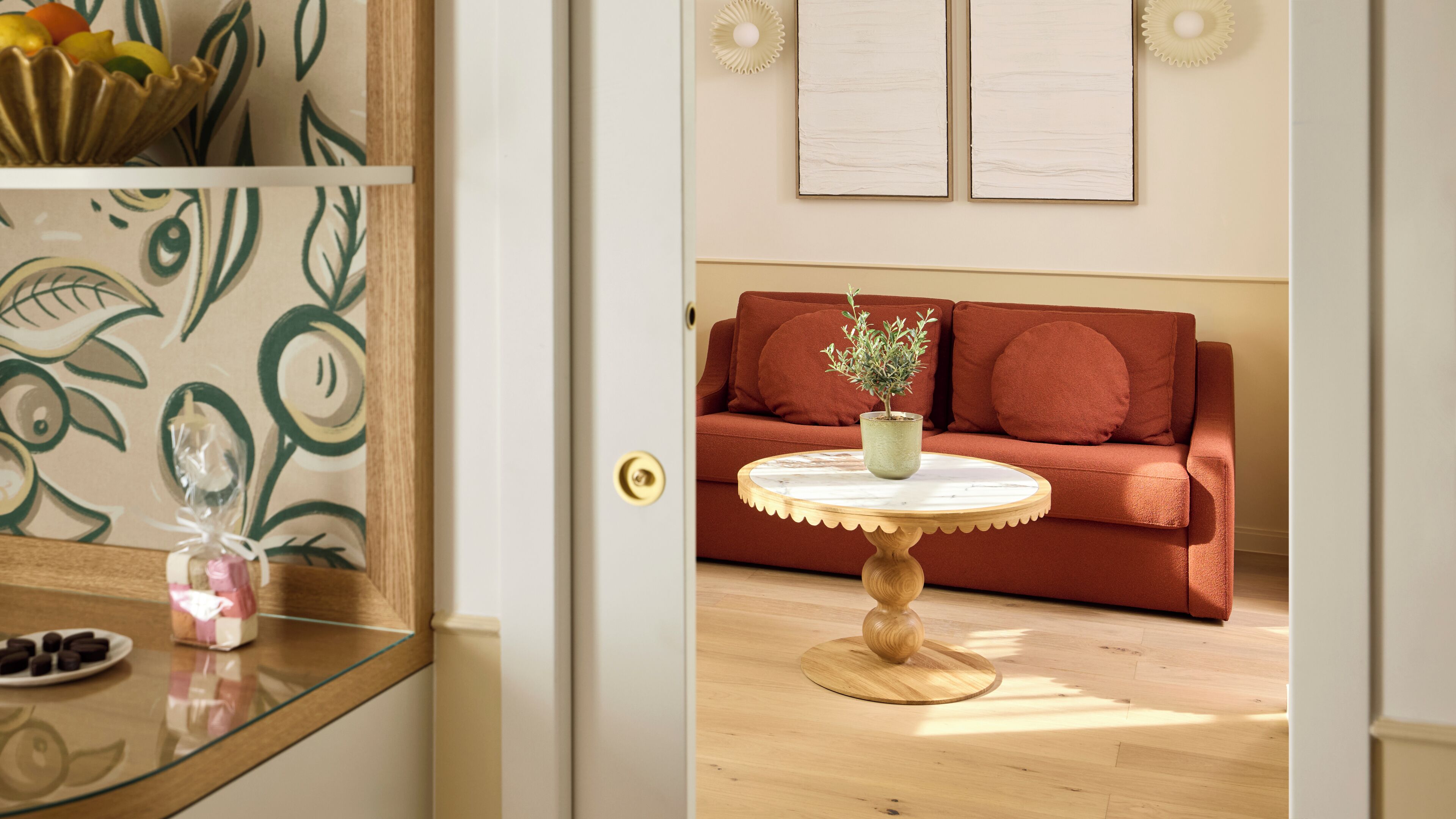 Una habitación con piso de madera, una mesa redonda con una planta verde, un sofá rojo y una pared con dos objetos enmarcados.
