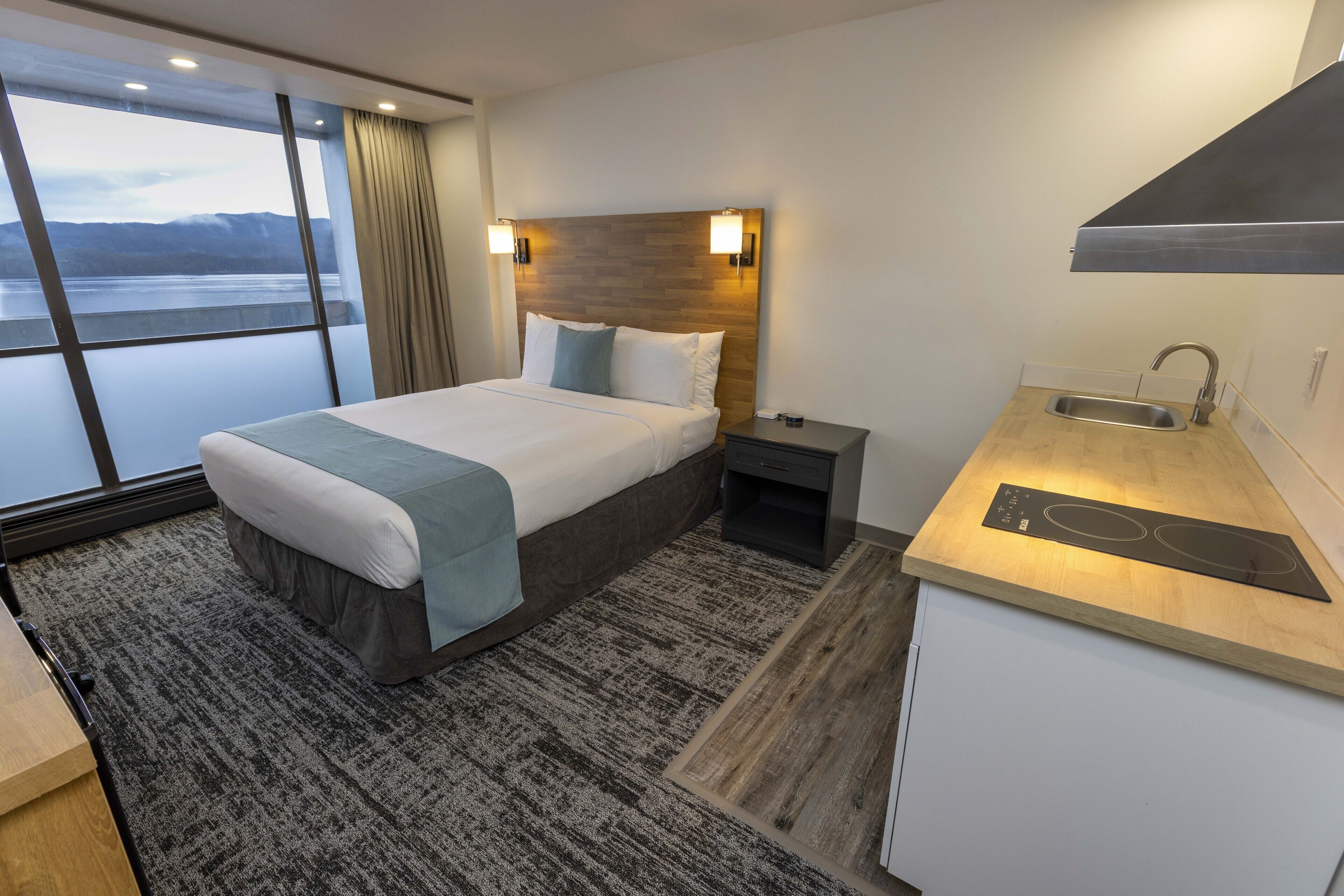 Habitación de hotel con una cama grande, una kitchenette pequeña, una ventana con vistas a un lago y un cabecero de madera.