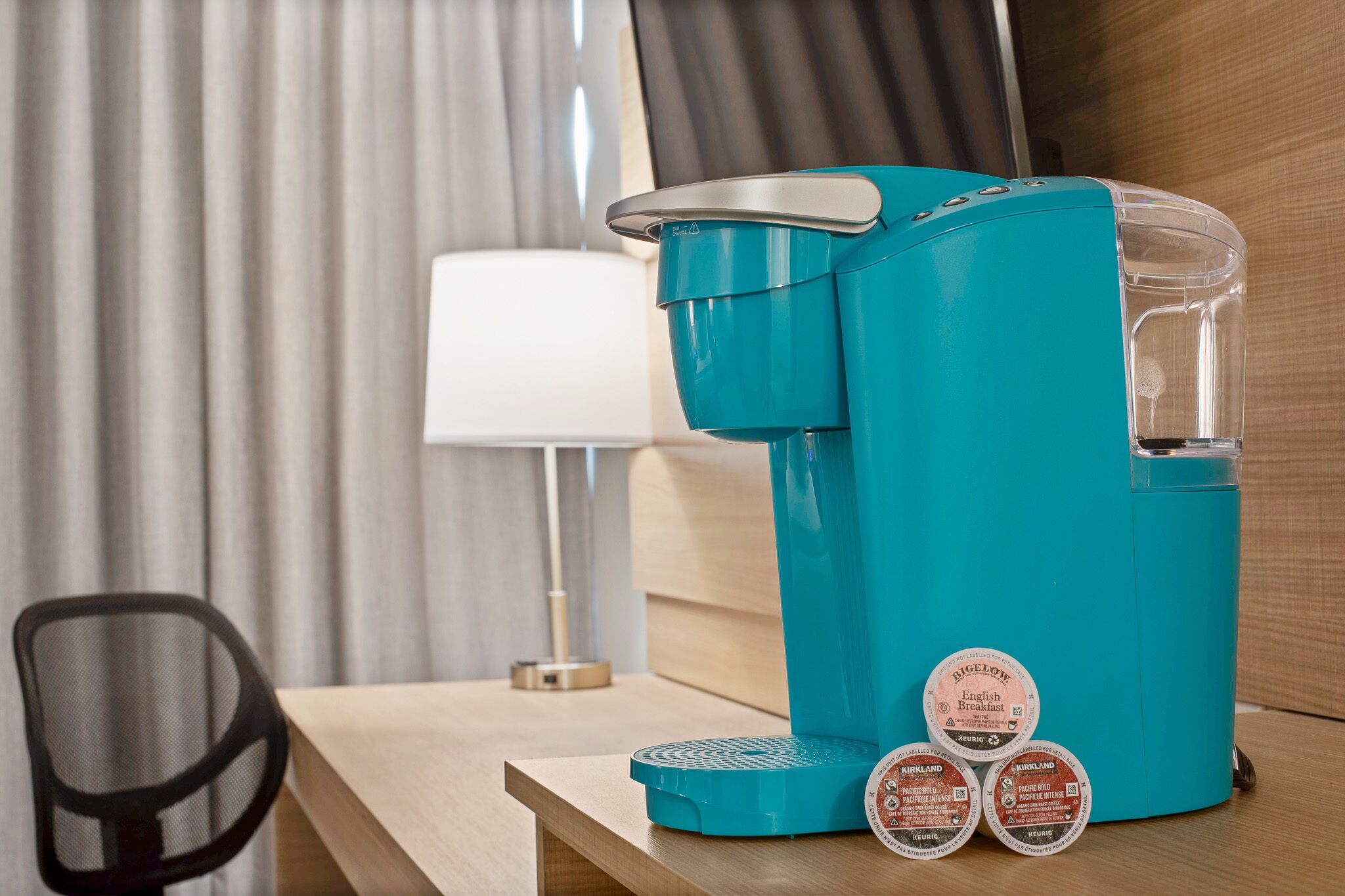 Eine türkisfarbene Kaffeemaschine mit transparentem Wasserbehälter und zwei Kaffee-Pods mit den Aufschriften 'BIGELOW English Breakfast' und 'KIRKLAND'.
