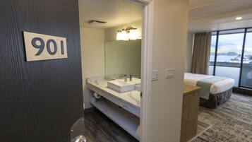 A hotel room with a bathroom featuring a sink, mirror, and towel rack. The room has a bed and a window with a view.