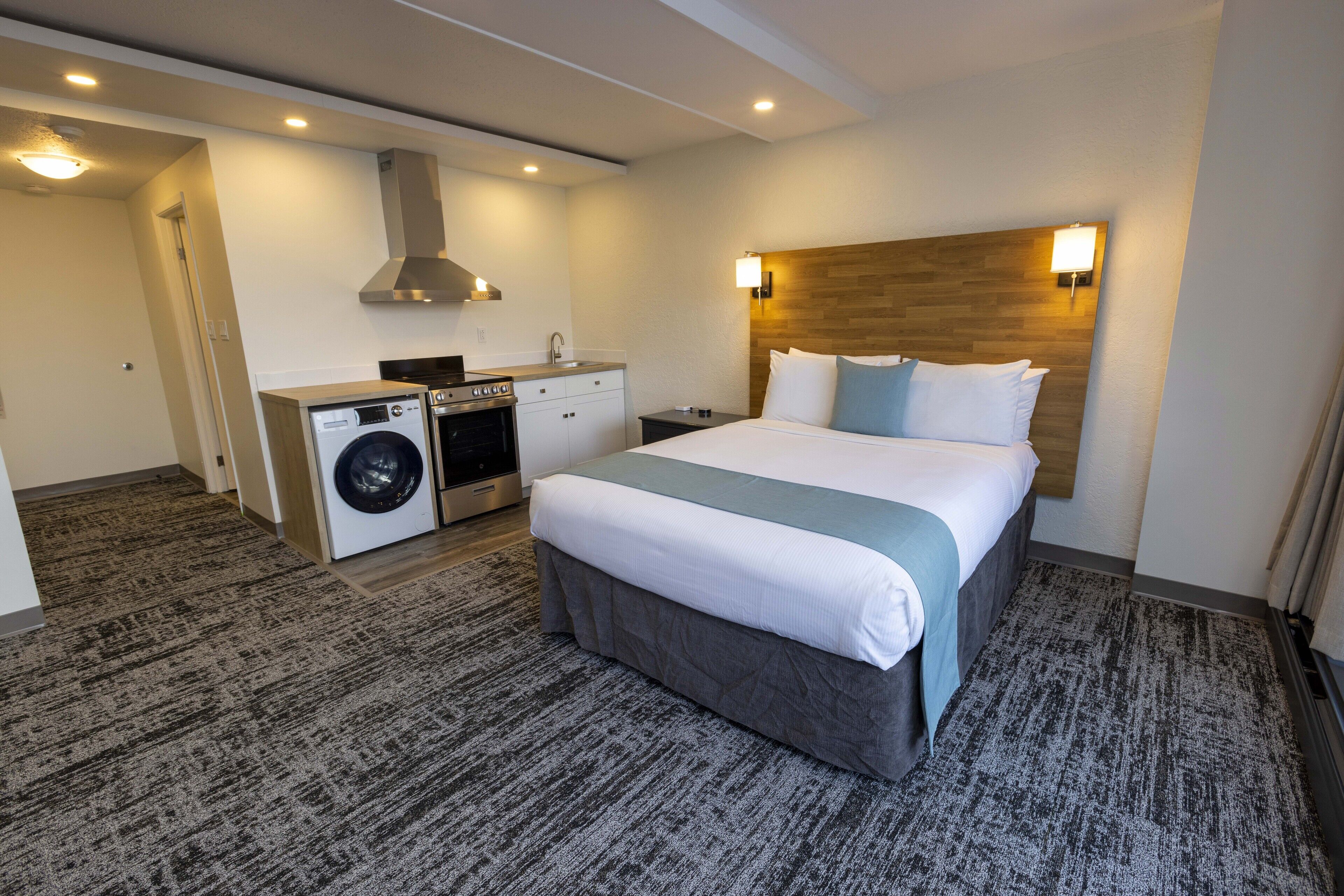 Habitación de hotel con cama, lavadora, kitchenette y cabecero de madera.