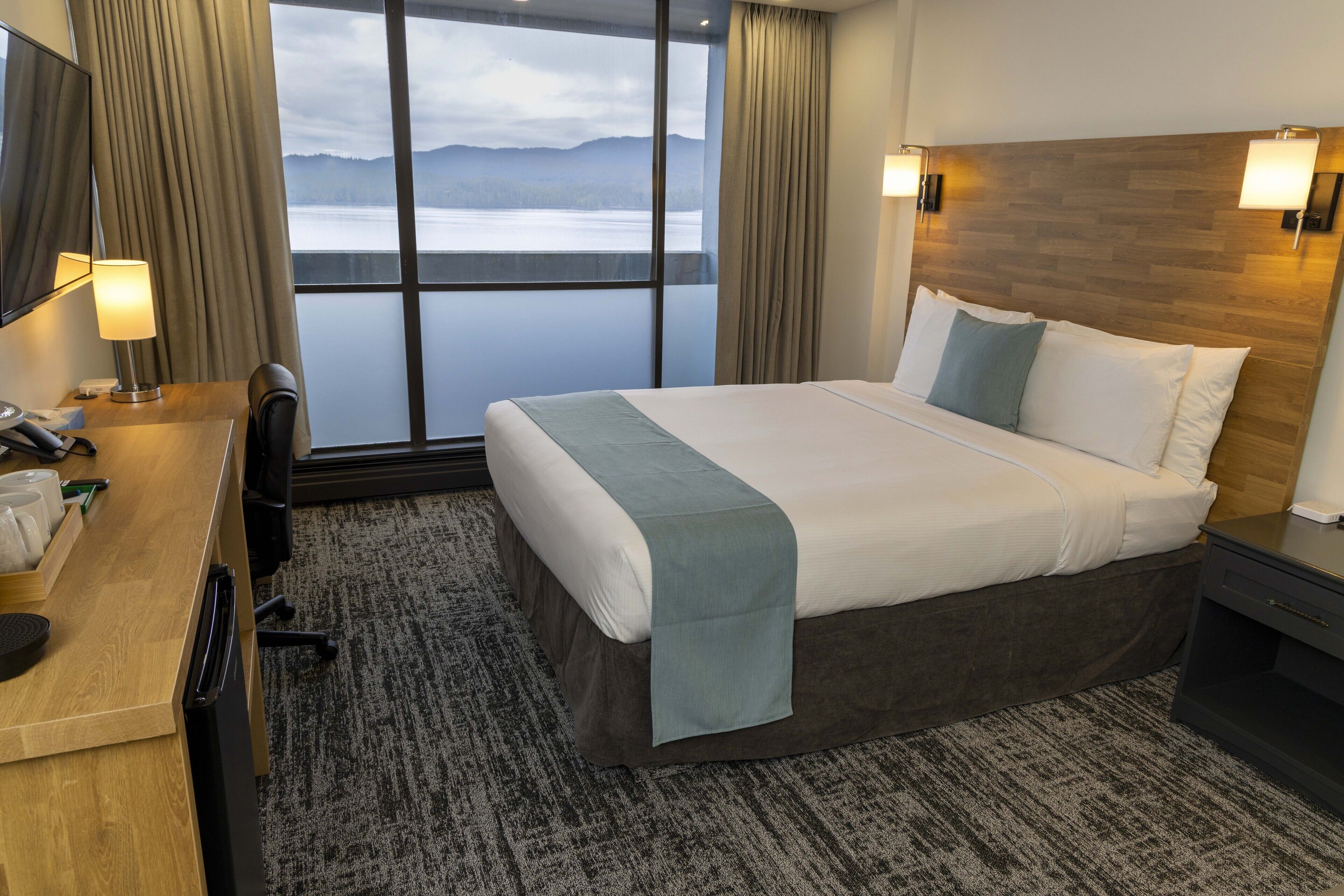 Habitación de hotel con una cama grande, un escritorio con lámpara, televisión y vista a un lago.