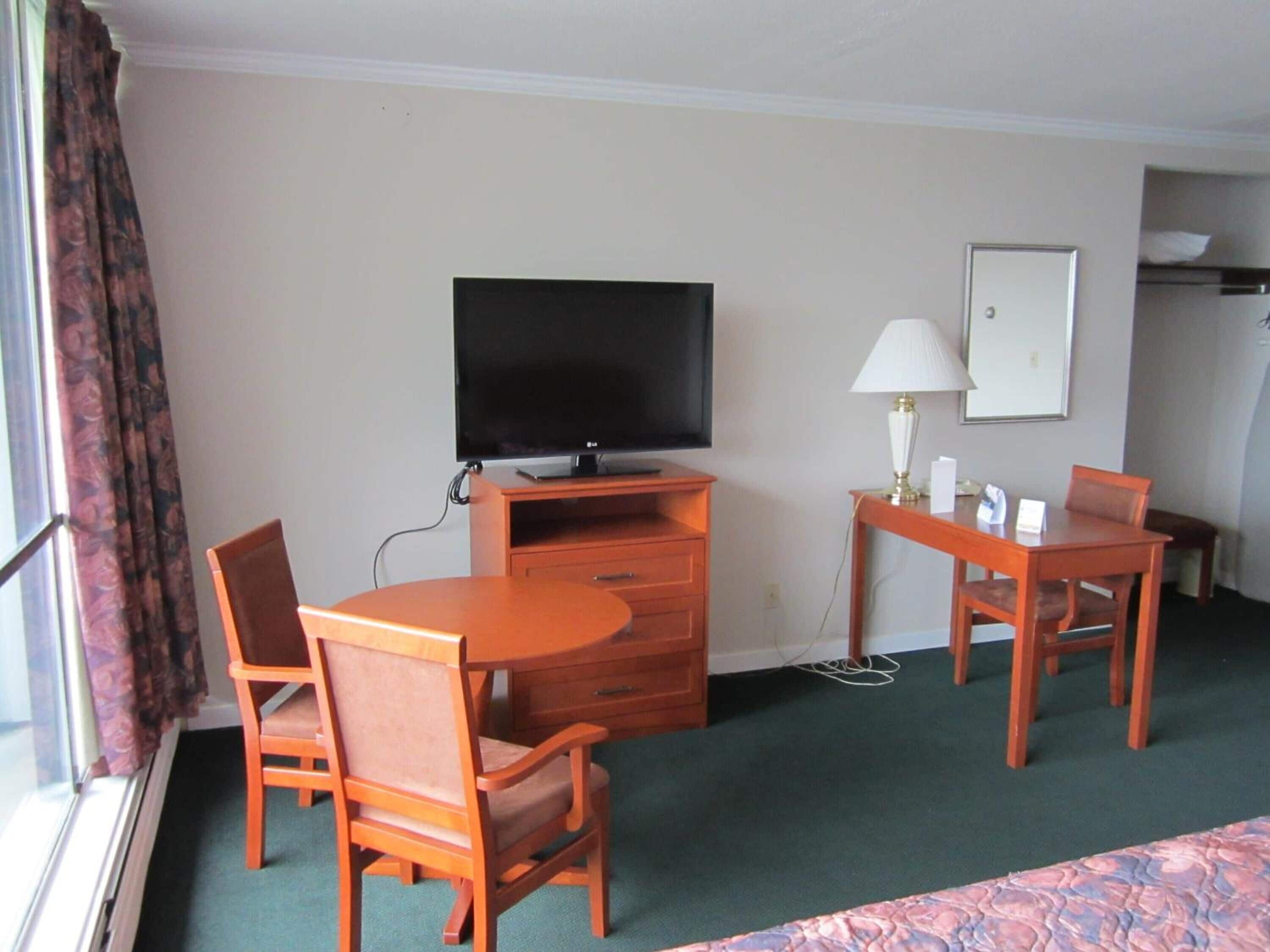 Habitación de hotel con televisor de pantalla plana, escritorio con lámpara, dos sillas, cama y ventana con cortinas.