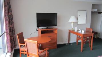 Quarto de hotel com televisão de tela plana, uma escrivaninha com abajur, duas cadeiras, uma cama e uma janela com cortinas.