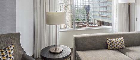 A modern hotel room with a grey sofa, a round side table, and a lamp. A window offers a view of a building and a palm tree.