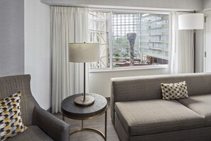 A modern hotel room with a grey sofa, a round side table, and a lamp. A window offers a view of a building and a palm tree.