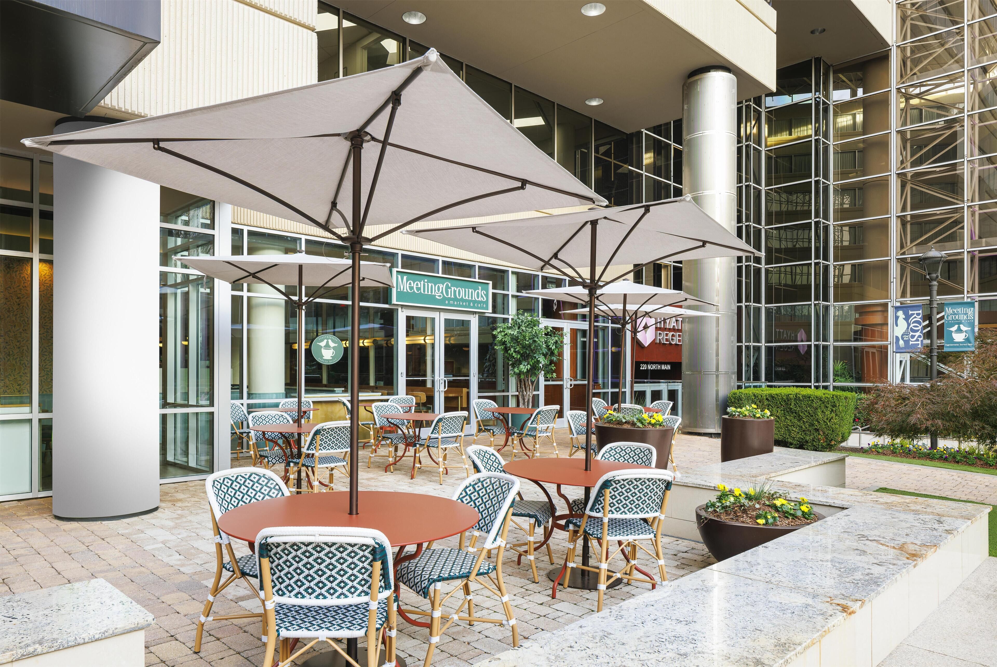 An outdoor seating area with tables and chairs, umbrellas, and a building with a sign that reads "Meeting Grounds."