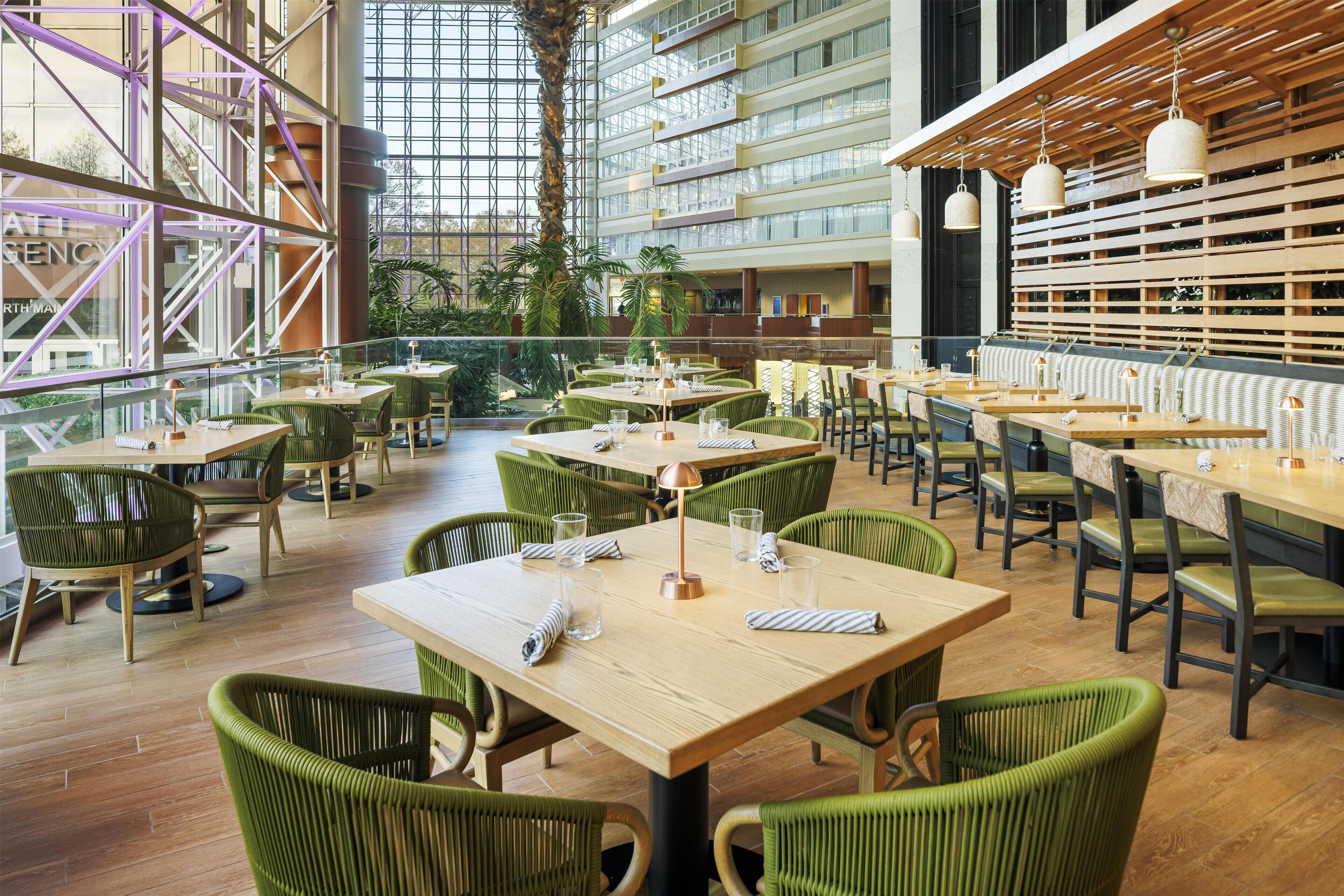 Un vaste espace intérieur aménagé en salle à manger, avec de hauts plafonds, de grandes fenêtres et des chaises recouvertes d’un tissu vert. Les tables sont disposées avec soin et chaque couvert est en place.