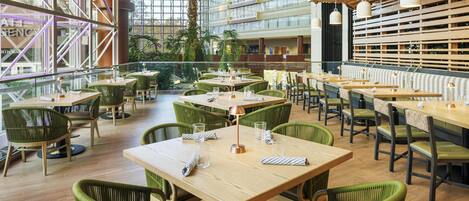 Un vaste espace intérieur aménagé en salle à manger, avec de hauts plafonds, de grandes fenêtres et des chaises recouvertes d’un tissu vert. Les tables sont disposées avec soin et chaque couvert est en place.