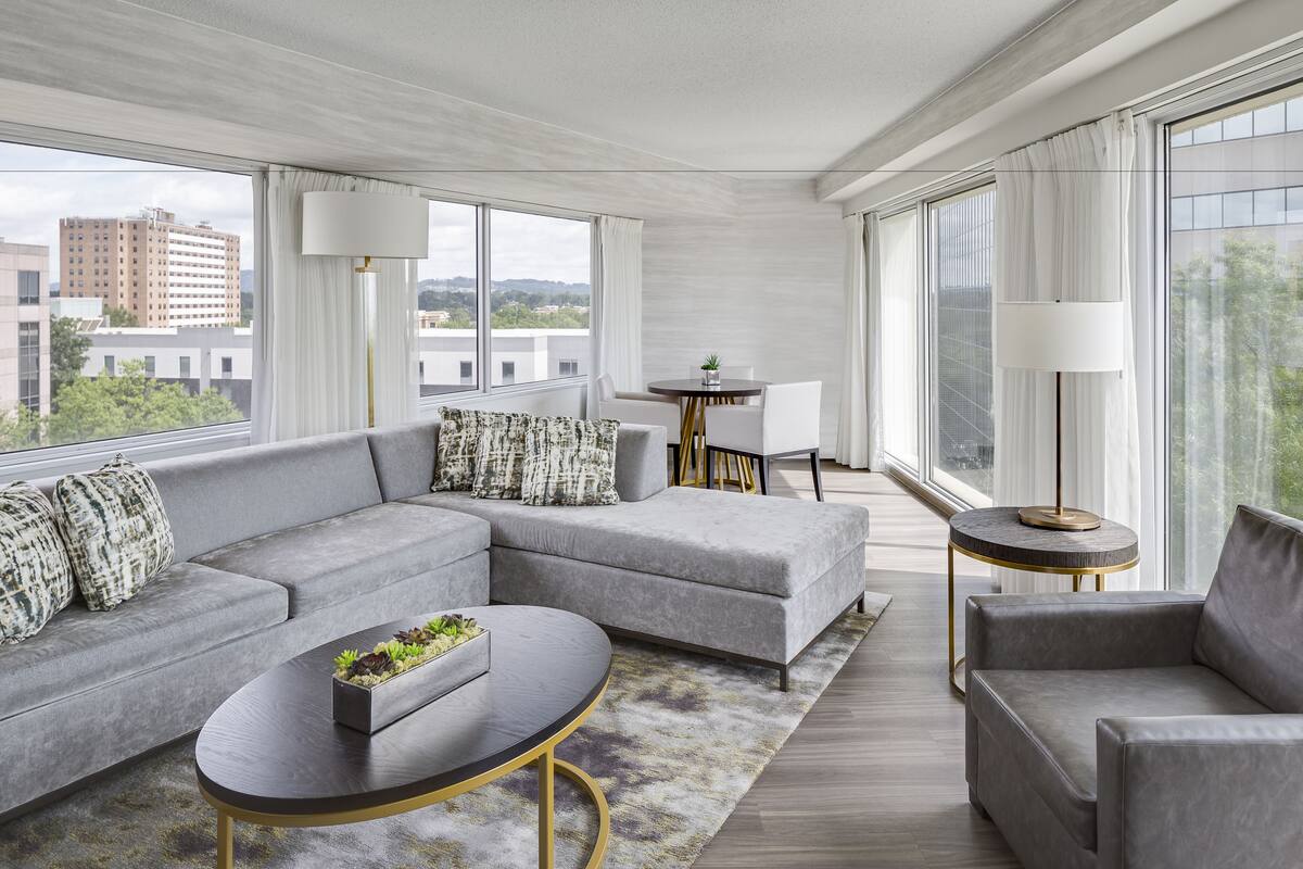 Ein modernes Wohnzimmer mit einem grauen Ecksofa, einem runden Couchtisch und einem kleinen Beistelltisch. Durch das Fenster ist ein Blick auf Gebäude möglich.