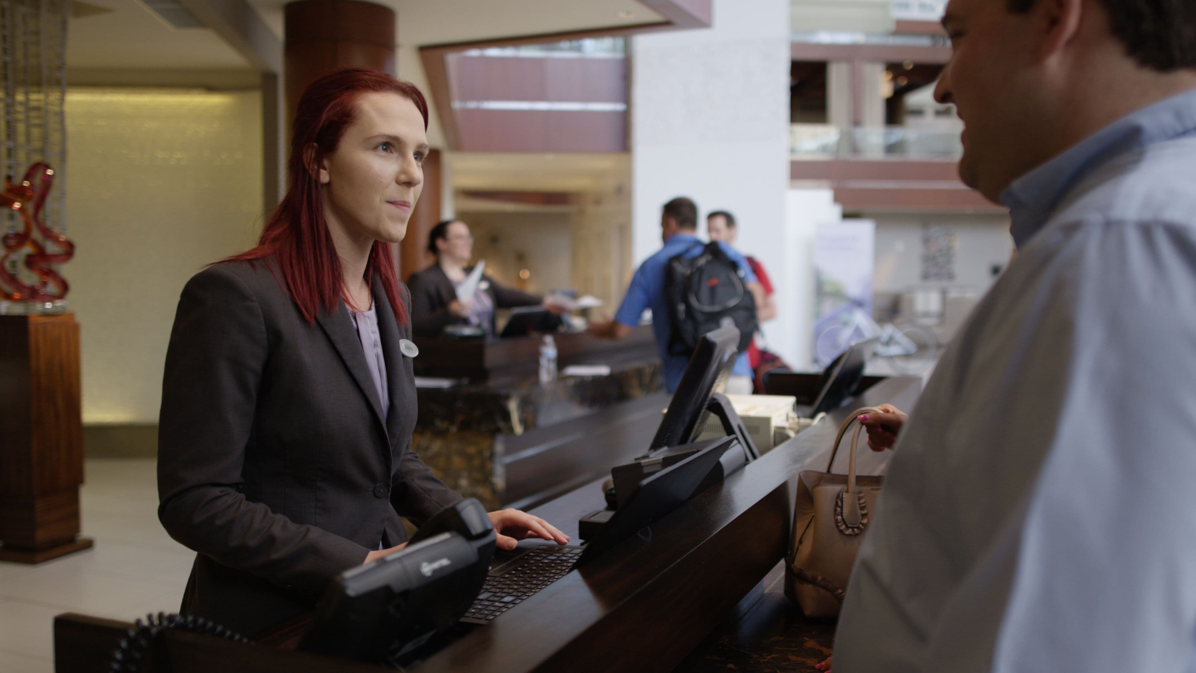 Une zone d'accueil avec un comptoir, un ordinateur et un téléphone. On aperçoit des personnes à l'arrière-plan, certaines assises et d'autres debout.