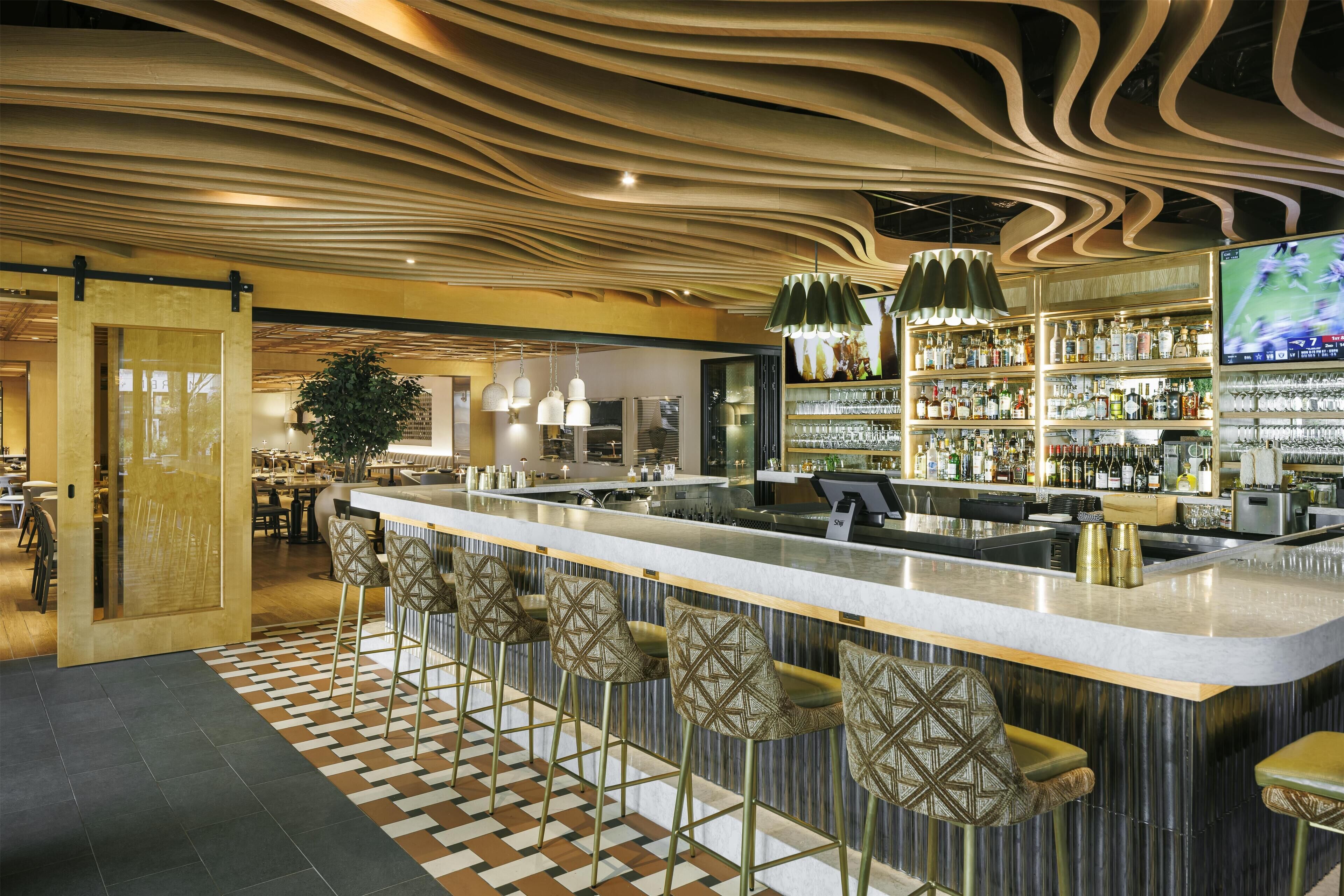 A bar area with a modern design, featuring a patterned floor, a long counter with stools, and a variety of bottles and glasses on shelves.