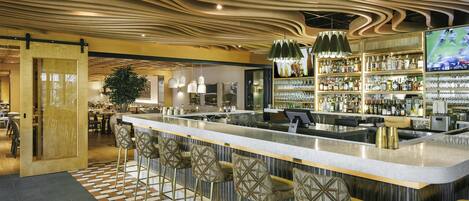 A bar area with a modern design, featuring a patterned floor, a long counter with stools, and a variety of bottles and glasses on shelves.