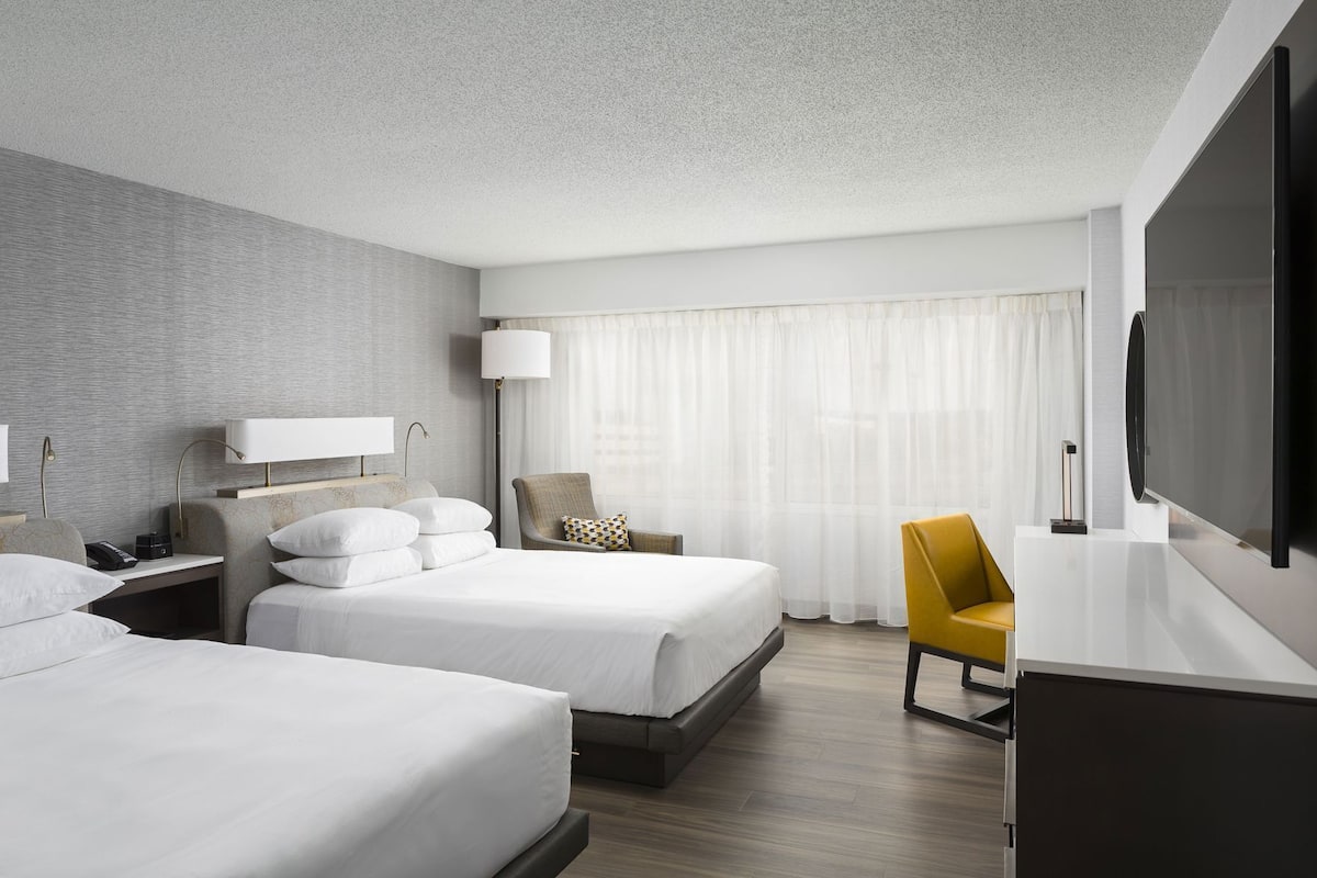 Ein Hotelzimmer mit zwei Betten, einem Schreibtisch, einem Stuhl, einem Fernseher und einem großen Fenster mit Vorhängen.