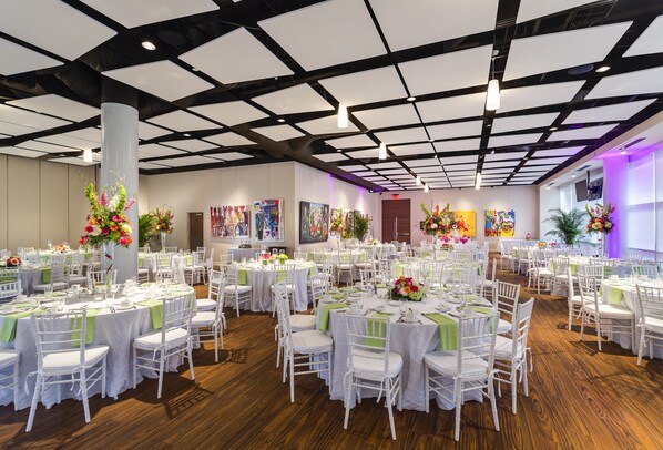 Un salón de banquetes con mesas redondas, sillas blancas y centros de mesa florales.