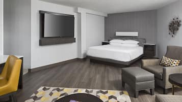 Une chambre d’hôtel moderne dotée d’un grand lit, d’un téléviseur à écran plat fixé au mur, d’un coin salon avec un fauteuil et un pouf, et d’une petite table.